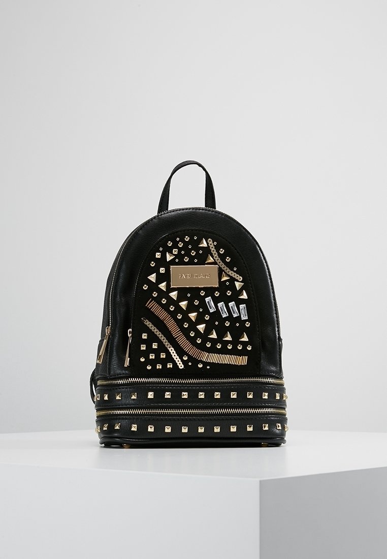 Mini sac à dos noir avec clous dorés, ornements géométriques, deux compartiments zippés et une petite poignée supérieure sur une surface blanche.