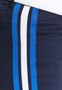 Marinblå byxor med blå och vita vertikala ränder längs sidorna, med en liten svart knapp med "Polo"-logotyp på fickan.