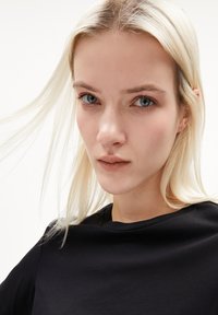 Zwart kortemouwen shirt met een ronde hals, gedragen door een persoon met steil, lichtblond haar en neutrale make-up, kijkend naar de camera.