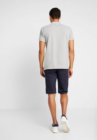 Uomo che si allontana indossando un polo a maniche corte grigio chiaro, pantaloni corti scuri a fantasia fino al ginocchio e sneakers bianche con accenti blu navy.
