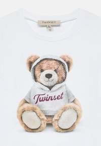 Fehér póló, amelyen egy medve grafika található, kapucnis pulóverrel díszítve, ezüst flitterekkel és bordó "Twinset" felirattal.