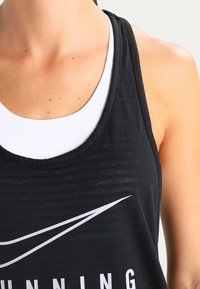 Musta urheilullinen tank top, jossa on syvä kaula-aukko, puuvillasekoitekangas ja valkoinen logo edessä. Kerroksittain valkoisen tanktopin päällä.