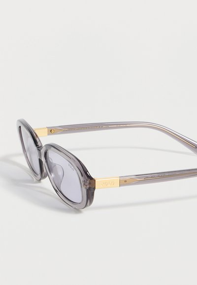 Γκρι διαφανή οβάλ γυαλιά ηλίου με χρυσές λεπτομέρειες και το λογότυπο "POLO" στις βραχίονες, εμφανίζονται σε λευκό φόντο.