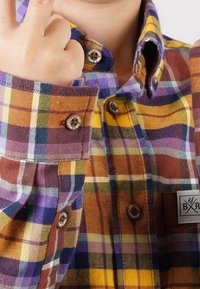 Chemise à carreaux avec des couleurs orange, jaune, violet et marron, dotée d'un col boutonné et d'un badge logo sur la poche avant. Tissu texturé.