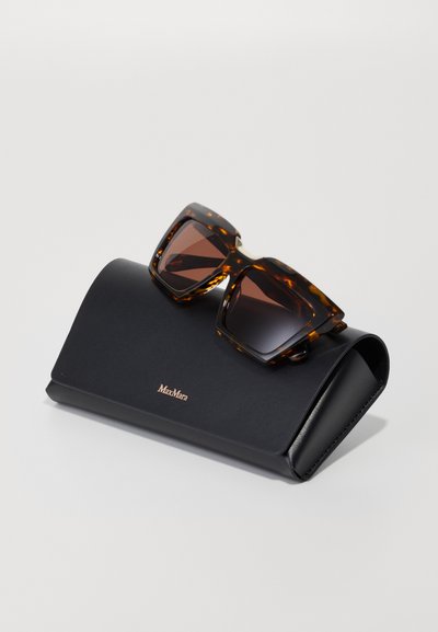 Des lunettes de soleil en écaille de tortue avec des verres sombres reposent sur un étui en cuir noir arborant un logo doré en relief à l'avant.