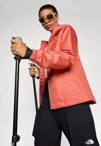 Coral waterproof jacket with elastic cuffs, black pants featuring cargo pockets, and ski poles; model wears sunglasses.