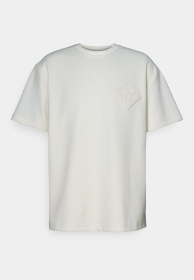 Lyhese varrukaga t-särk heledas puuvillas, millel on vasakul rinnal tekstuuriga rombikujuline pitsat ja väljaulatuv disain.