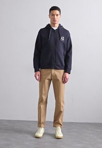 Hanorac negru cu fermoar, cu buzunar frontal, cămașă gri dedesubt, pantaloni chino bej și adidași de culoare deschisă. Țesătură fină cu detalii de logo.