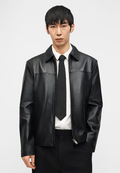 Άνδρας με μαύρο δερμάτινο μπουφάν πάνω από λευκό πουκάμισο και μαύρη γραβάτα, που στέκεται μπροστά σε απλό λευκό φόντο.