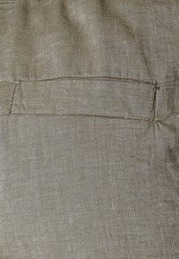 Beżowy materiał lniany z delikatną strukturą, z szwem tylnej kieszeni. Materiał ma lekki, organiczny wygląd.