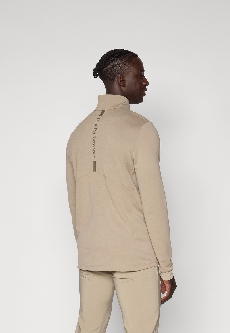 Beige langermet topp med høy krage og detaljert søm på baksiden. Har en subtil logo trykket vertikalt langs ryggraden. Glatt, lett stoff.