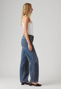 Laiade säärega sinised teksapüksid kõrge vöökohaga, millel on klassikaline viies taskuga disain ja kontrastsed õmblused. Kandmisel valge top.