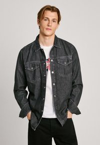 Zwarte denimjas met knoopsluiting, twee borstzakken en stikdetails. Draag het over een witte graphic t-shirt met ronde hals.