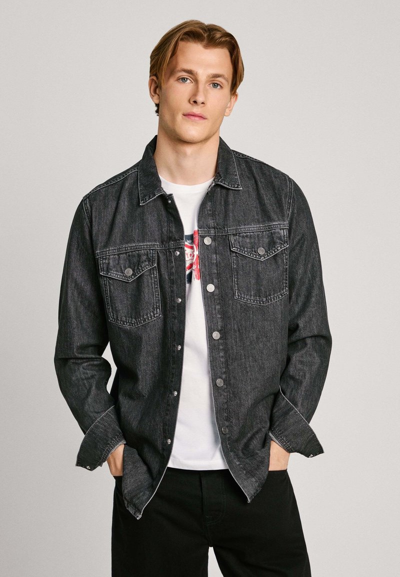 Zwarte denimjas met knoopsluiting, twee borstzakken en stikdetails. Draag het over een witte graphic t-shirt met ronde hals.