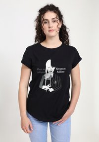 Mladá žena s kudrnatými vlasy nosí černé tričko s rozděleným obrazem postavy z Addamsovy rodiny, která drží knihu, text zní: "Jednou Addams, navždy Addams."