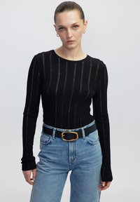 Schwarzes Langarmoberteil mit vertikalen strukturierten Streifen, kombiniert mit hochgeschnittenen hellblauen Jeans und einem schwarzen Gürtel mit einer goldenen Schnalle.