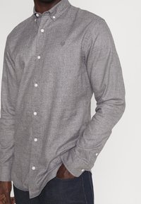 Koszula na guziki w szarym melanżowym materiale, z kołnierzykiem, długimi rękawami i białymi guzikami. Zawiera małe logo na piersi.