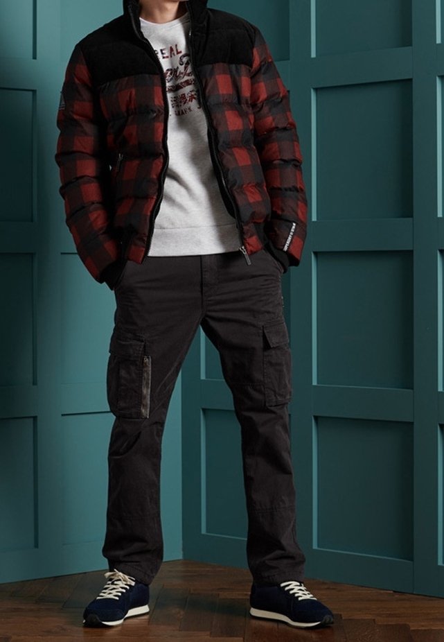 Homme portant une doudoune à carreaux rouges et noirs, un sweat-shirt blanc à motifs, un pantalon cargo noir et des baskets bleu marine, debout devant un mur à panneaux bleu sarcelle.