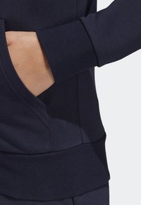 Main insérée dans la poche avant d'un sweat-shirt bleu marine foncé avec des poignets et une ceinture côtelés.