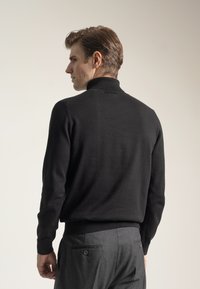 Hombre vistiendo un suéter negro de cuello alto y pantalones grises, de pie y mirando hacia un lado frente a un fondo claro y liso.