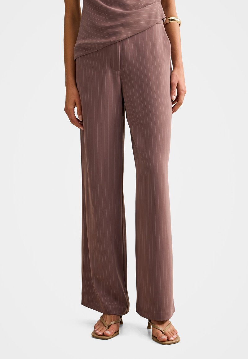 Vrouw die mauve wijde pijp broek met krijtstrepen draagt, beige sandalen met hakken en een gouden armband om haar rechterpols tegen een witte achtergrond.