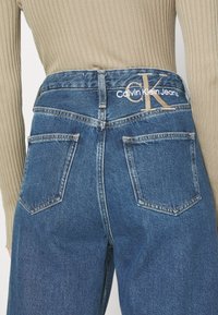 Denim hlače znamke Calvin Klein v srednje modri barvi z visokim pasom, ki vključujejo zadnje žepe in logotipne detajle v beli ter bež niti.