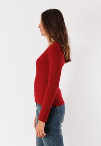 Top rosso a maniche lunghe a coste con silhouette aderente e scollatura a V. Abbinato a jeans in denim blu, creando un look informale.
