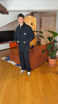 Črna platnena jakna s sprednjimi žepi, črne hlače in srebrni športni copati. Soba ima leseno tla in rastlino blizu rjavega kordurovske kavča.