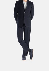 Costume marine à deux boutons, coupe ajustée, poches détaillées, et une chemise de couleur claire ; associé à des chaussures en daim bleu marine et des chaussettes foncées.