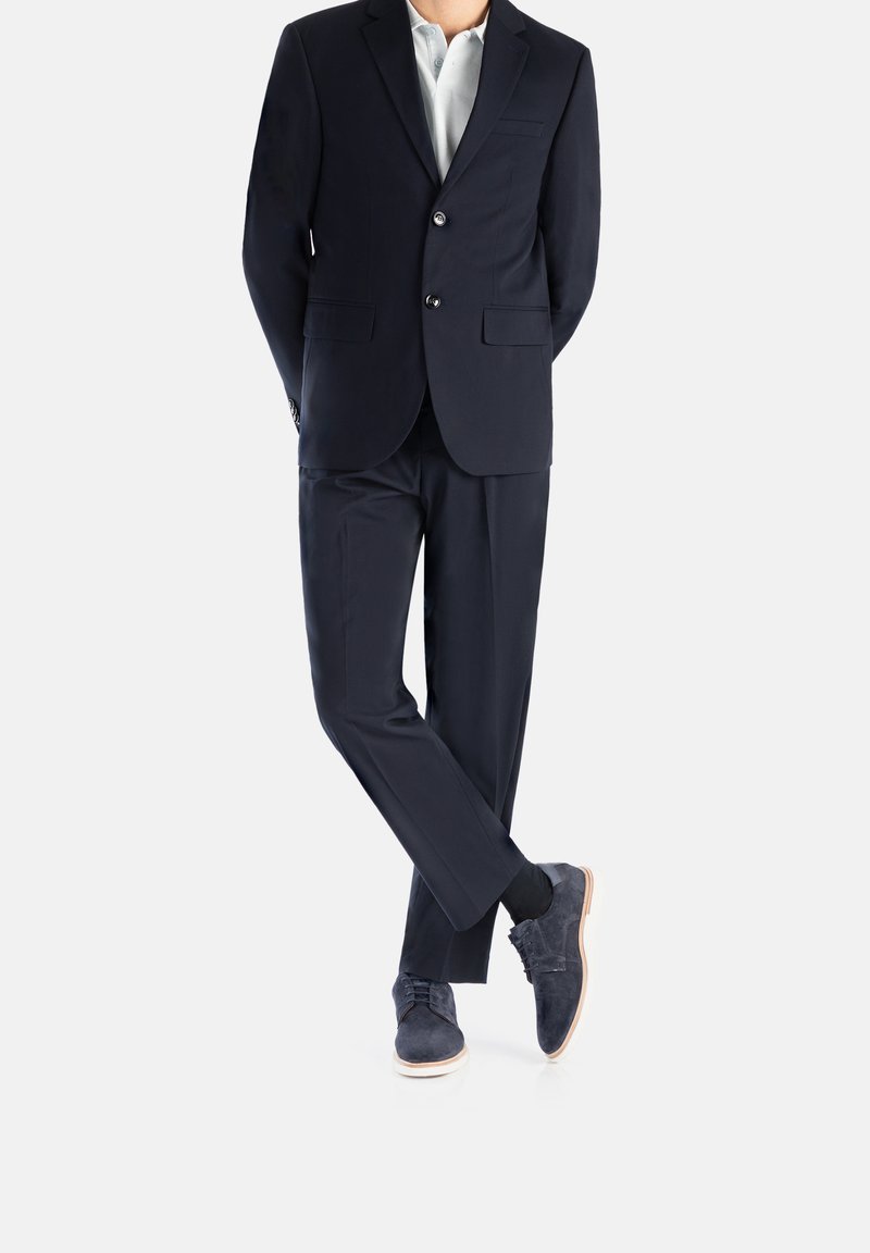 Costume marine à deux boutons, coupe ajustée, poches détaillées, et une chemise de couleur claire ; associé à des chaussures en daim bleu marine et des chaussettes foncées.