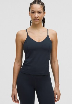 Frau mit geflochtenem Haar trägt ein eng anliegendes schwarzes ärmelloses Oberteil und schwarze Leggings, steht vor einem schlichten hellen Hintergrund.