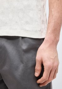 Κοντινό ενός χεριού που ακουμπάει στο πλάι, με λευκό πουκάμισο με υφή και το λογότυπο «HUGO» κοντά στο τελείωμα και γκρι παντελόνι.