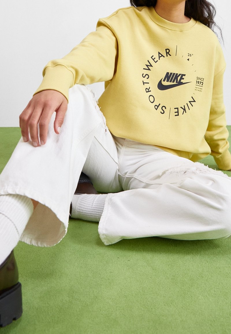 Žlutá mikina s černým logem Nike a nápisem, doplněná o roztrhané bílé široké kalhoty a černé boty. Silné žebrované manžety a kulatý výstřih.