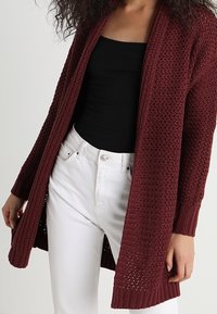 Burgundy stickad cardigan med öppen framsida, långa ärmar, ribbade ärmslut och en texturerad mönster. Bärs över en svart topp och vita byxor.