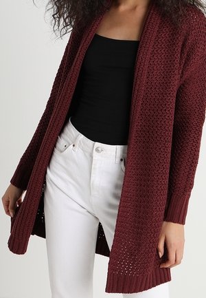 Cardigan en tricot bourgogne à ouverture sur le devant, à manches longues, avec des poignets côtelés et un motif texturé. Porté sur un haut noir et un pantalon blanc.