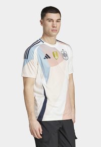 Camisa de futebol de manga curta em cores pastel com padrões geométricos em azul, rosa e branco; possui um emblema da FIFA e um patch da Espanha.