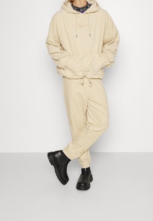 Hoodie in corduroy beige con tasca frontale e cordoncino, abbinato a pantaloni con cordoncino coordinati; stivaletti neri alla caviglia con suola spessa.