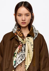Bufanda de seda con estampados mixtos, que presenta diseños de leopardo, florales y patrones en tonos beige, marrón y negro. Atada al cuello, superpuesta a una chaqueta marrón.