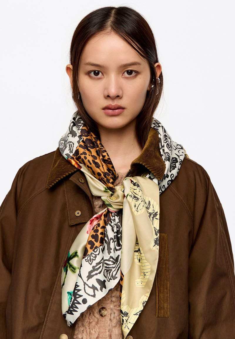 Bufanda de seda con estampados mixtos, que presenta diseños de leopardo, florales y patrones en tonos beige, marrón y negro. Atada al cuello, superpuesta a una chaqueta marrón.