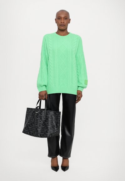 Helle grüne Strickpullover mit übergroßen Ärmeln, kombiniert mit schwarzen Kunstlederhosen und einer gemusterten schwarzen Tragetasche.