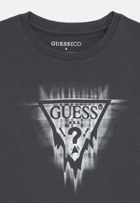 Mørk grå crewneck skjorte med et forvrengt hvitt og svart GUESS-logo og spørsmålstegn grafikk på brystet samt størrelse 8 etikett inni kragen.