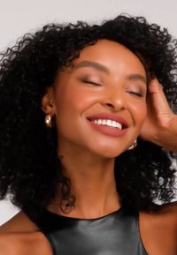 Kiharatukkainen nainen hymyilee suljettu silmin, kullanväriset renkorenkaat korvakorissaan ja musta hihaton yläosa, käsi pään päällä lepäämässä.