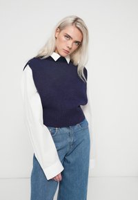 Sweater sem mangas azul marinho feito de tecido macio, apresentando uma bainha canelada e detalhes em botão nos ombros, usado sobre uma camisa branca de colarinho.