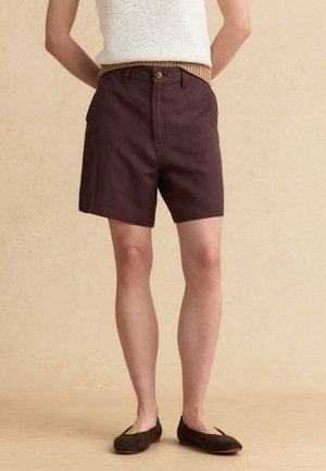 Persoon draagt donkerbruine, op maat gemaakte korte broek, een mouwloos gebreid topje en donkerbruine platte schoenen, staand tegen een neutrale achtergrond.