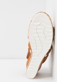 Sandale avec une semelle texturée blanche, des brides marron et une bordure tressée multicolore, positionnée verticalement contre une surface blanche.