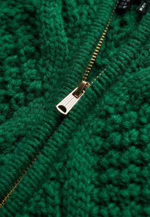 Suéter verde de punto con un patrón texturizado y un cierre con cremallera metálica dorada. La lana parece gruesa y acogedora.