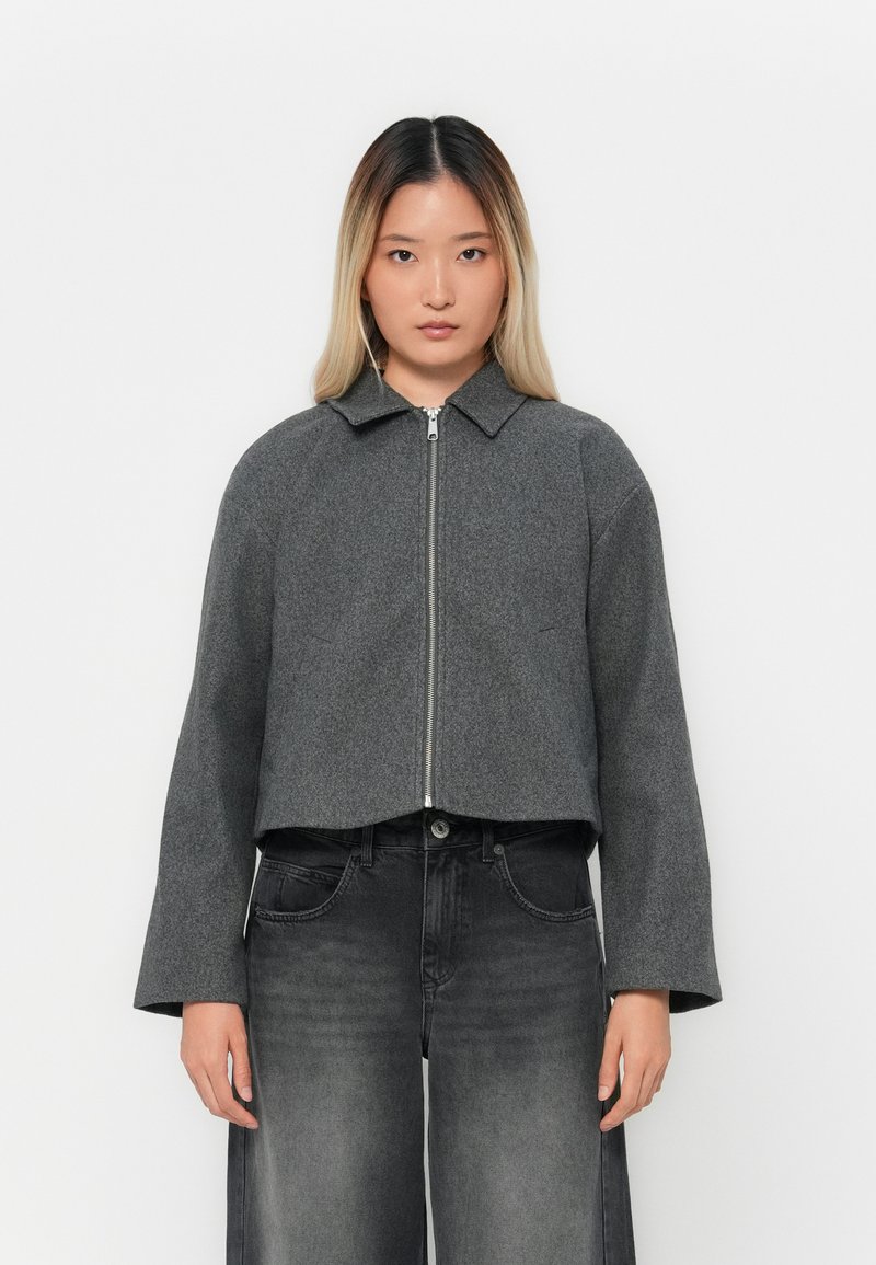 Giacca grigia corta realizzata in misto lana, con chiusura frontale a zip, maniche larghe e colletto classico. Abbinata a jeans grigio scuro.