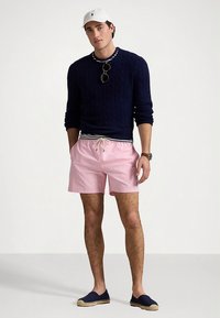 Μπλε πουλόβερ με πλεκτό σχέδιο, ροζ μαγιό σορτς με κορδόνι, μπλε εσπαντρίγιες και λευκό καπέλο. Αξεσουάρ περιλαμβάνουν γυαλιά ηλίου και ρολόι χειρός.