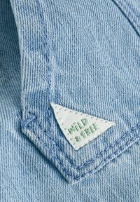 Tissu en denim bleu clair avec une étiquette triangulaire blanche portant l'inscription "WILD & FREE" cousue dans un coin, mettant en avant un design de vêtement décontracté.