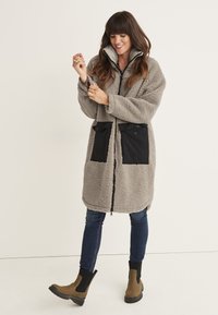 Gray plush coat with a zip front, black fabric pockets, and a high collar. Worn over blue jeans and brown ankle boots. Smooth texture.